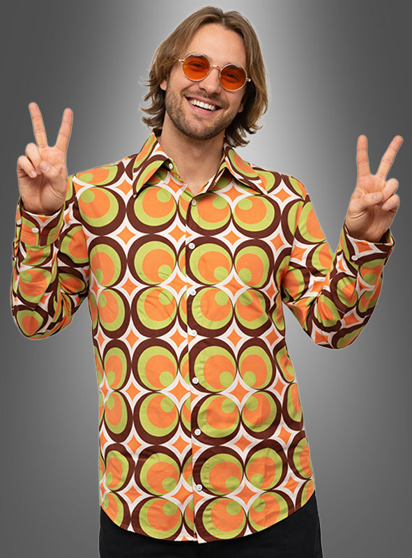Ein Mann, der ein hochwertiges, waschbares 70er-Jahre-Retro-Hemd namens „Nick“ mit einem orangefarbenen, grünen und braunen Retro-Muster trägt und dazu eine passende orangefarbene Sonnenbrille, lächelt und macht mit beiden Händen vor einem grauen Hintergrund das Peace-Zeichen – perfekt für jede 70er-Jahre-Party.