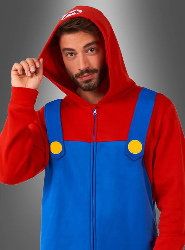 Ein Mann, der den „Super Mario Overall für Erwachsene“ trägt – bestehend aus einem roten Kapuzenpullover, einem blauen Latzhosen-Design und gelben Knöpfen, die vom lizenzierten Super-Mario-Kostüm inspiriert sind –, steht vor einem grauen Hintergrund.