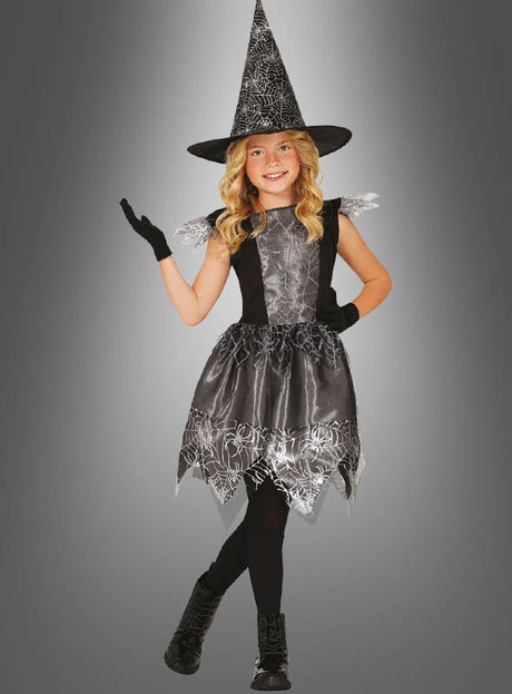 Ein junges Mädchen, das ein Hexenkostüm für Kinder zu Halloween trägt – mit einem hohen Hut, schwarzen Handschuhen und Stiefeln –, steht auf einem Bein vor einem grauen Hintergrund mit Farbverlauf.