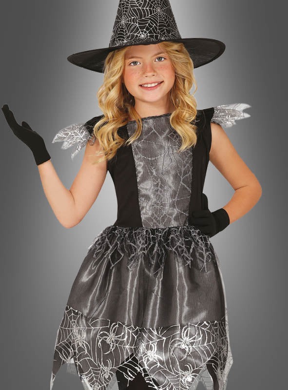 Ein junges Mädchen trägt das schwarze Hexenkostüm für Kinder zu Halloween mit silbernen Spinnennetz-Details und einem passenden Hexenhut und posiert mit erhobenem Arm.