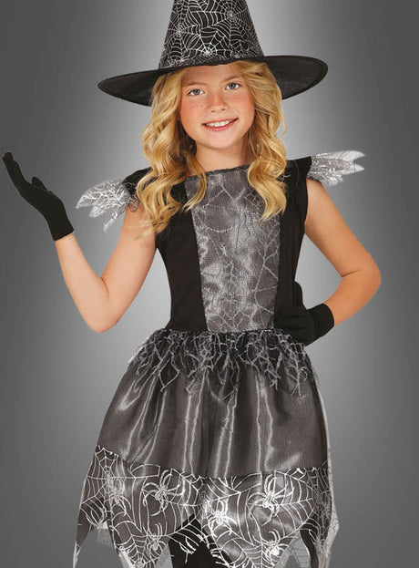 Ein junges Mädchen trägt das schwarze Hexenkostüm für Kinder zu Halloween mit silbernen Spinnennetz-Details und einem passenden Hexenhut und posiert mit erhobenem Arm.