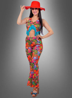Eine Frau, die das Hippie-Style-2-Teiler-Set „Yuna“ im Bauchtanz-Boho-Stil trägt – ein farbenfrohes, bauchfreies Boho-Set mit einem leuchtend orangefarbenen Hut, einer Sonnenbrille, einem farbenfrohen Neckholder-Top mit Blumenmuster und blauen Fransen, einer passenden Schlaghose sowie Plateausandalen – vor einem grauen Hintergrund mit Farbverlauf und verkörpert damit perfekt den Hippie-Kostüm-Look für Damen, die den neuesten Hippie-Style-Trend mitmachen.