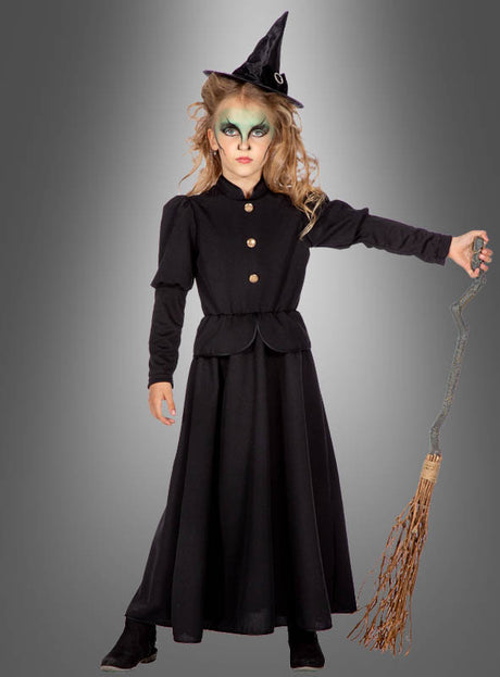 Ein Kind, das ein „Klassisches Hexenkleid Kinder“ trägt – ein langes schwarzes Hexenkostüm –, komplett mit spitzem Hut, auffälligem Make-up und einem Besen in der Hand, steht vor einem grauen Hintergrund mit Farbverlauf.