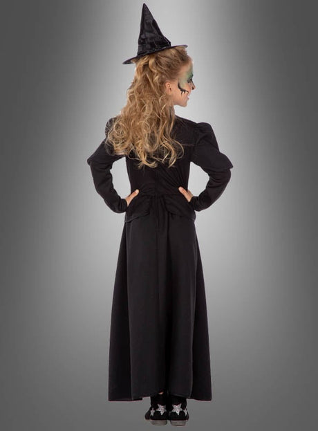 Ein Kind, das ein klassisches Hexenkleid für Kinder trägt und einen spitzen Hexenhut auf dem Kopf hat, steht mit den Händen in die Hüften gestemmt vor einem grauen Hintergrund und blickt von der Kamera weg.