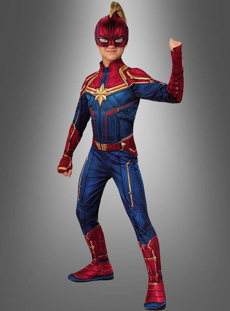 Ein Kind in einem „Captain Marvel für Mädchen“-Kostüm posiert mit erhobener Faust. Es trägt einen rot-blau-goldenen Anzug, komplett mit Maske und Stiefeln, vor einem grauen Hintergrund – das ideale Marvel-Superheldinnen-Kostüm für jede Karnevalsfeier.