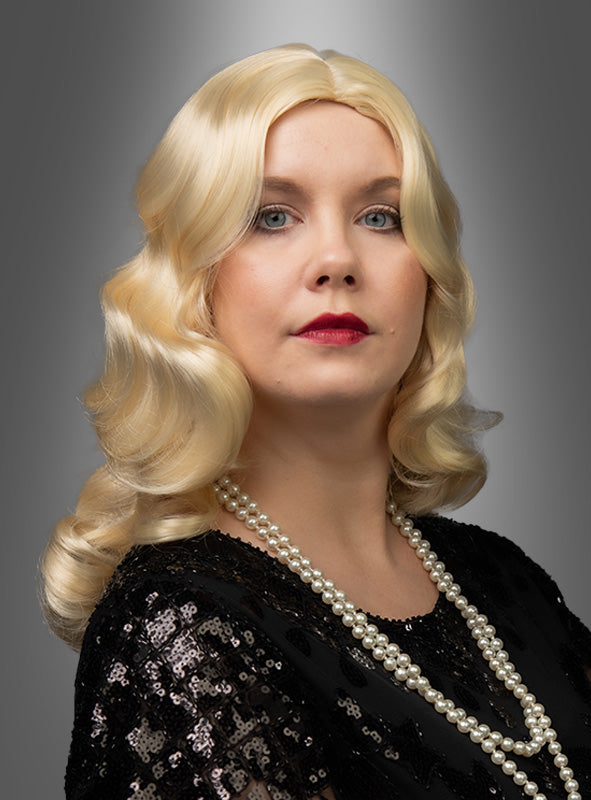 Eine Frau mit welligem blondem Haar, das mit einer „Luxus 20er Premium Wasserwelle Perücke Elli“ frisiert ist, trägt ein schwarzes Top mit Pailletten und eine Perlenkette und posiert vor einem grauen Hintergrund mit Farbverlauf.