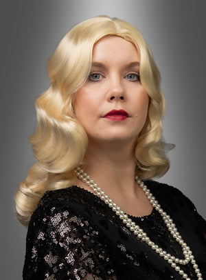 Eine Frau mit welligem blondem Haar, das mit einer „Luxus 20er Premium Wasserwelle Perücke Elli“ frisiert ist, trägt ein schwarzes Top mit Pailletten und eine Perlenkette und posiert vor einem grauen Hintergrund mit Farbverlauf.