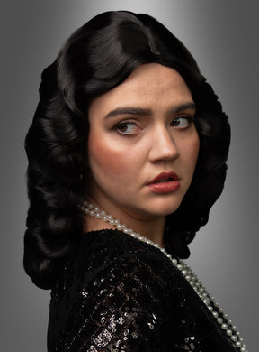 Eine Frau mit welligem, dunklem Haar im Vintage-Look trägt ein schwarzes Pailletten-Oberteil und eine Perlenkette und blickt vor einem grauen Hintergrund mit Farbverlauf über die Schulter. Ihr glamouröses Erscheinungsbild erinnert an die Eleganz der „Luxus 20er Premium Wasserwelle Perücke Elli“, die sich durch klassische Wasserwellen auszeichnet.
