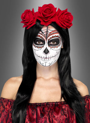 Eine Person mit langen schwarzen Haaren, die einen roten Haarschmuck mit Blumenmuster zum Tag der Toten sowie ein rotes Kleid trägt, hat ihr Gesicht im Stil eines Zuckerschädels zum Tag der Toten mit schwarzen, weißen und roten Details bemalt, wodurch ein authentischer Zuckerschädel-Kostüm-Look entsteht.