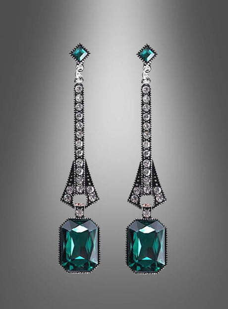 Ein Paar „Elegante Ohrringe für Damen, Silber“ mit langem, silbernem Tropfendesign, verziert mit grünen Edelsteinen und kleinen klaren Steinen, die symmetrisch angeordnet sind, dargestellt vor einem grauen Hintergrund mit Farbverlauf.