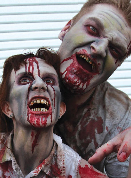 Zwei als Zombies verkleidete Personen mit blutverschmierten Gesichtern, grauem Make-up und gelben Zähnen posieren vor einem weißen Hintergrund und präsentieren dabei beeindruckende SFX-Filmqualität. Ihr furchterregendes Aussehen wird durch die „Zombie-Zahnfarbe Filmqualität Deluxe“ zum Leben erweckt, was dieses Kostüm zum perfekten Halloween-Kostüm für jede gruselige Veranstaltung macht.
