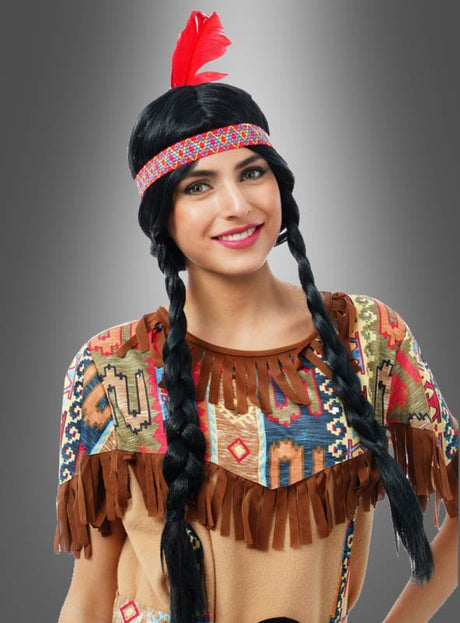 Eine Person trägt die „Indianerperücke für Erwachsene“ – eine geflochtene schwarze Perücke – sowie ein Stirnband mit einer roten Feder und ein gemustertes Kostüm mit Fransen und posiert vor einem grauen Hintergrund mit Farbverlauf.