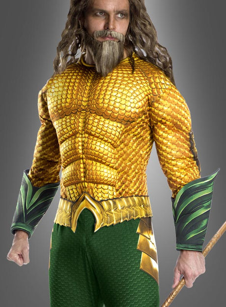 Eine Person, die das Aquaman-Herrenkostüm trägt – mit einem goldenen Schuppenmuster, grünen Akzenten und einem Dreizack in der Hand – steht vor einem grauen Hintergrund.