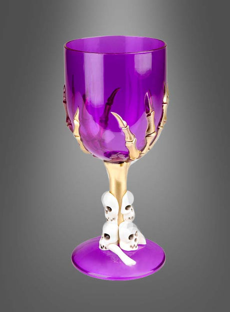 Das Halloween-Weinglas mit Skeletthand (17 cm) besteht aus einem violetten Kelch, an dessen Schale eine goldene Skeletthand festhält, während kleine weiße Totenköpfe den Stiel zieren – ein auffälliges Halloween-Dekostück, das vor einem grauen Hintergrund fotografiert wurde.