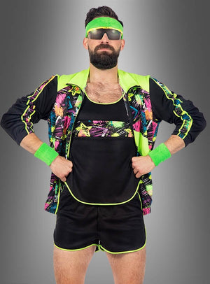 Ein Mann, der einen neongrünen und schwarzen „Retro Neon Aerobic Fitness Anzug Herren“ – einen kurzen Trainingsanzug im 80er-Jahre-Stil – trägt, dazu eine Sonnenbrille, ein Stirnband und Schweißbänder, steht mit den Händen in den Hüften vor einem grauen Hintergrund.