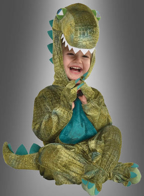 Ein Kind sitzt lächelnd vor einem grauen Hintergrund und trägt das „Dino-Kostüm für Kleinkinder“, ein grünes Kostüm mit einer Kapuze in Form eines Dinosaurierkopfes – perfekt für den Kinderfasching.