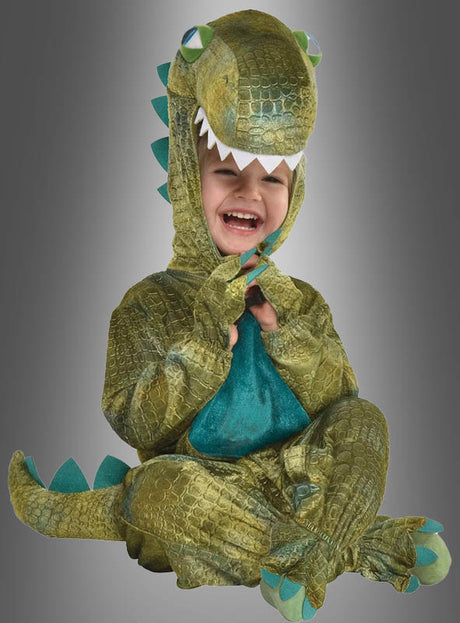 Ein Kind sitzt lächelnd vor einem grauen Hintergrund und trägt das „Dino-Kostüm für Kleinkinder“, ein grünes Kostüm mit einer Kapuze in Form eines Dinosaurierkopfes – perfekt für den Kinderfasching.