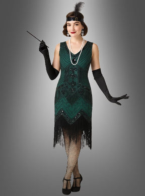 Eine Frau präsentiert das „20er-Jahre-Kleid Shirley“ mit Accessoires in Grün – bestehend aus einem grünen Charleston-Kleid, schwarzen Handschuhen, Netzstrümpfen, Perlen, einem Haarreif mit Federn und einer Zigarettenspitze –, und fängt so vor einem grauen Hintergrund die Essenz der klassischen Accessoires der „Roaring Twenties“ ein.