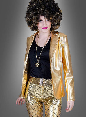Eine Person trägt die „Glänzende Disco Jacke Damen“ in Gold, glänzende goldene Shorts, Netzstrümpfe, ein schwarzes Shirt, eine goldene Halskette und eine schwarze Lockenperücke und steht vor einem schlichten grauen Hintergrund.
