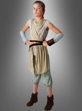 Eine junge Person nimmt mit den Händen in den Hüften eine entschlossene Pose ein, gekleidet in ein beige- und hellgraues Outfit, vor einem grauen Studiohintergrund – ideal für alle, die auf der Suche nach dem authentischen Star Wars-Rey-Kinderkostüm sind.