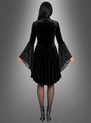 Eine Frau, die das Gothic-Kleid „Lucy mit Spitze“ aus schwarzem Samt mit ausgestellten Trompetenärmeln trägt und dazu Stiefeletten kombiniert hat, steht vor einem grauen Hintergrund mit Farbverlauf und wendet der Kamera den Rücken zu.