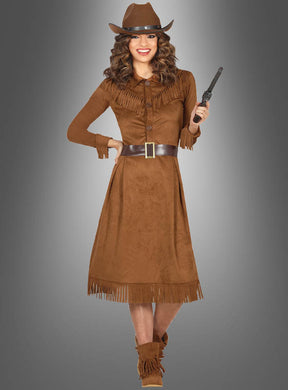 Eine Frau, gekleidet in ein braunes, mit Fransen verziertes Cowgirl-Westernkleid für Damen, trägt einen Hut und hält einen Spielzeugrevolver in der Hand; sie steht vor einem grauen Hintergrund mit Farbverlauf.