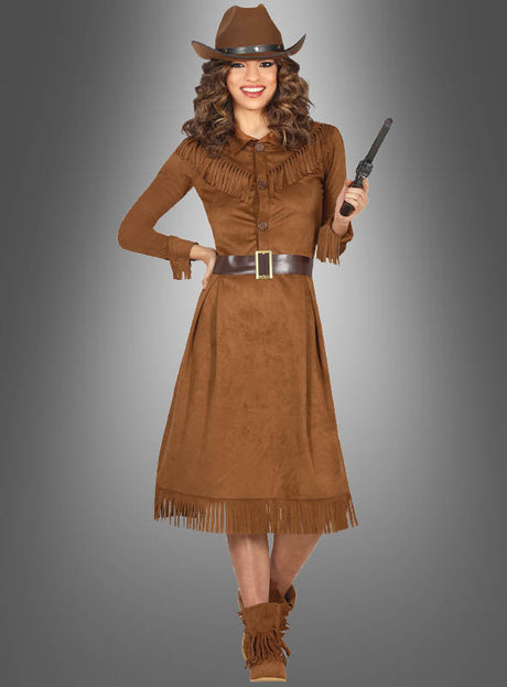 Eine Frau, gekleidet in ein braunes, mit Fransen verziertes Cowgirl-Westernkleid für Damen, trägt einen Hut und hält einen Spielzeugrevolver in der Hand; sie steht vor einem grauen Hintergrund mit Farbverlauf.