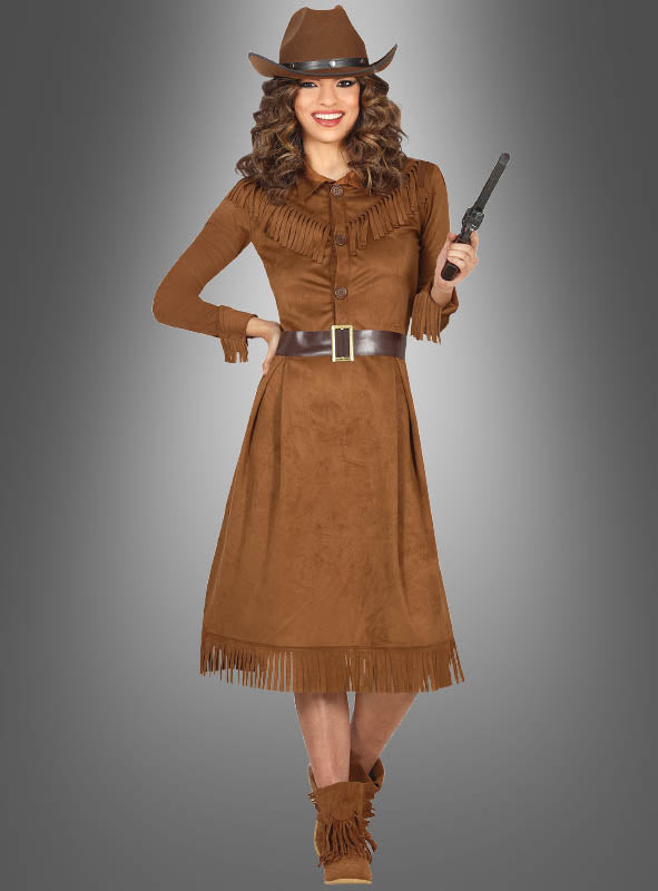 Eine Frau, gekleidet in ein braunes, mit Fransen verziertes Cowgirl-Westernkleid für Damen, trägt einen Hut und hält einen Spielzeugrevolver in der Hand; sie steht vor einem grauen Hintergrund mit Farbverlauf.