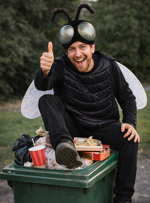 Eine Person in einem Fliegenkostüm, die wie eine lästige Mücke auf einer Mülltonne im Freien sitzt, ist von Lebensmittelverpackungen umgeben, streckt den Daumen nach oben und lächelt in die Kamera.