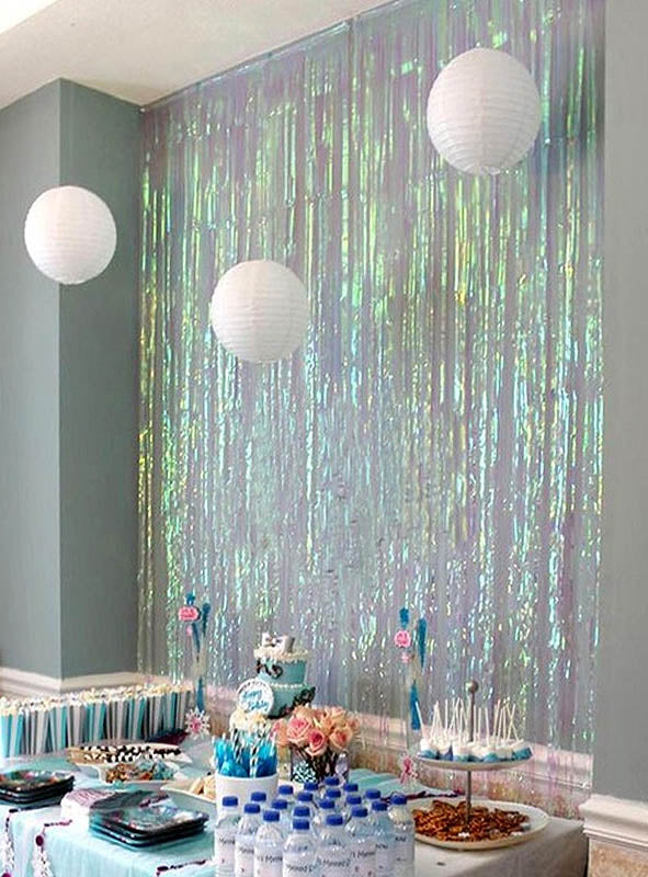 Vor einem schimmernden Lametta-Vorhang (200–250 cm) als Partydekoration steht ein Partytisch mit Snacks und Getränken. Über dem Tisch hängen drei weiße Papierlaternen, die das festliche Bild abrunden.