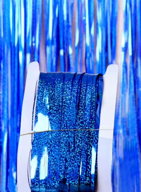 Eine Rolle glitzerndes blaues Band liegt in einem weißen Spender, der vor einem „Lametta Vorhang 200–250 cm Party Deko“ aus schimmerndem blauem Lametta platziert ist – perfekt für eine stilvolle Partydekoration.