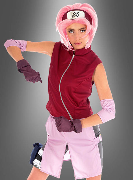 Eine Person mit einer rosa Perücke und einer roten Weste posiert im Sakura-Kostüm aus „Naruto Shippuden“, komplett mit Handschuhen, einem Stirnband mit dem Blatt-Symbol und hellrosa Shorts, die an den ikonischen Look von Sakura Haruno erinnern.