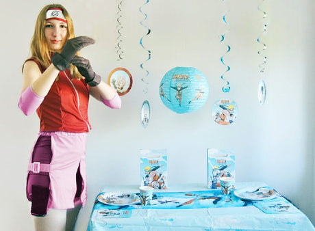 Eine Person in einem Sakura-Kostüm aus „Naruto Shippuden“ posiert neben einem geschmückten Tisch mit Partyzubehör zum Thema „Naruto“, blauen Hängedekorationen und einem hellen Hintergrund und erweckt so die lebendige Welt des Anime zum Leben.