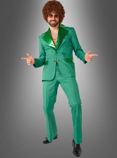 Ein Mann, der einen grünen Disco-Anzug mit Paillettenkragen trägt, ergänzt durch eine Afro-Perücke, eine Sonnenbrille und eine Goldkette, nimmt eine Pose ein, während er mit beiden Fingern zeigt – ein perfekter 70er-Jahre-Anzug für jede Karnevalskostümparty.