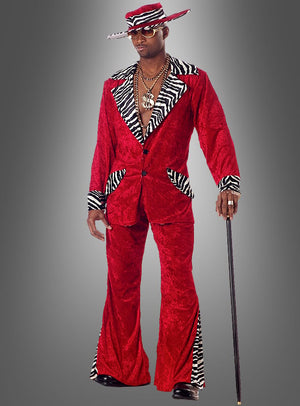 Eine Person, die das Zuhälter-Outfit „Rot mit Zebramuster“ trägt – bestehend aus einem roten Samtanzug mit Zebra-Print-Besatz, einem dazu passenden Hut mit breiter Krempe, goldenen Halsketten und einer Sonnenbrille sowie einem Spazierstock –, steht vor einem grauen Hintergrund.