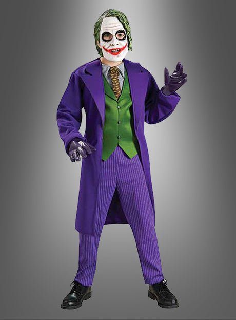 Ein Kind, das das Joker-Kostüm „Kinder Deluxe“ aus der Batman-Reihe trägt – komplett mit lila Mantel, grüner Weste, Nadelstreifenhose, Handschuhen und der typischen weißen Gesichtsbemalung mit roten Lippen und schwarzem Augen-Make-up –, steht vor einem grauen Hintergrund.
