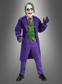 Ein Kind, das das Joker-Kostüm „Kinder Deluxe“ aus der Batman-Reihe trägt – komplett mit lila Mantel, grüner Weste, Nadelstreifenhose, Handschuhen und der typischen weißen Gesichtsbemalung mit roten Lippen und schwarzem Augen-Make-up –, steht vor einem grauen Hintergrund.