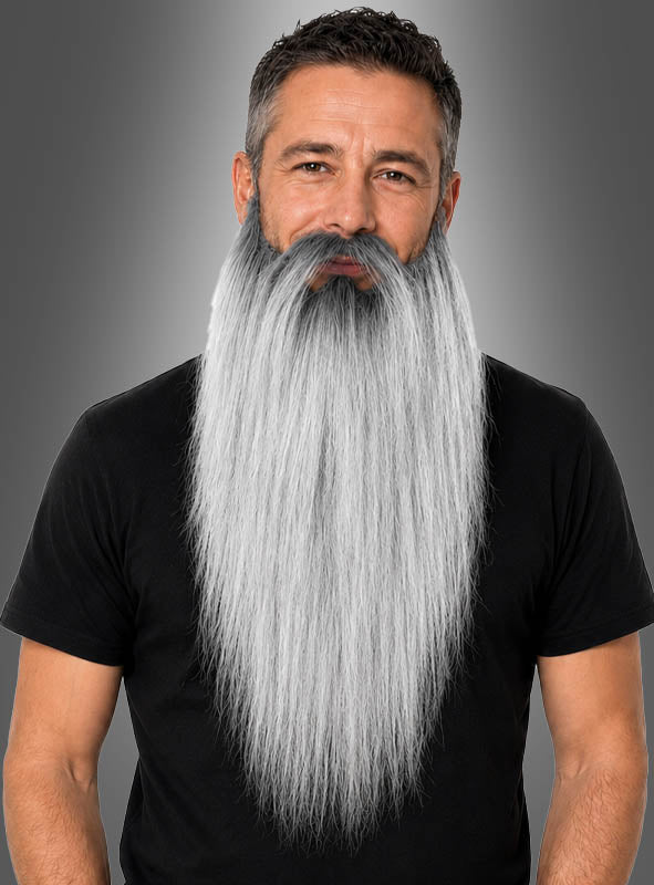 Ein Mann mit kurzen dunklen Haaren und einem ungewöhnlich langen, glatten, grauen „Langer Bart für Zauberer & Zwerge“, der an den Bart eines Zauberers erinnert, trägt ein schwarzes T-Shirt und steht vor einem grauen Hintergrund mit Farbverlauf.