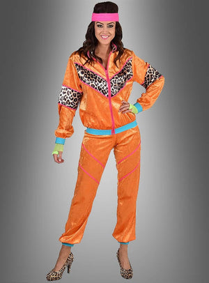 Eine Frau in einem XXL-Trainingsanzug im 80er-Jahre-Look in leuchtendem Orange mit Leopardenmuster-Details, kombiniert mit einem rosa Stirnband und passenden High Heels mit Leopardenmuster, steht lächelnd vor einem grauen Hintergrund – eine ideale Wahl für eine authentische 80er-Jahre-Party oder als auffälliges 80er-Jahre-Outfit.