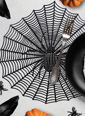 Halloween-Tischsets in verschiedenen Designs unter einer Gabel und einem schwarzen Teller, mit gruseligen Motiven wie Plastikspinnen, schwarzen Federn und kleinen orangefarbenen Kürbissen auf einem weißen Tisch.