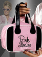 Eine Hand hält die Handtasche „Pink Ladies Grease“ – eine rosa-schwarze Tasche mit der Aufschrift „Pink Ladies“ –, die perfekt für „Grease“-Fans ist, während im Hintergrund jemand in einer passenden Jacke und mit einer rosa Sonnenbrille steht.