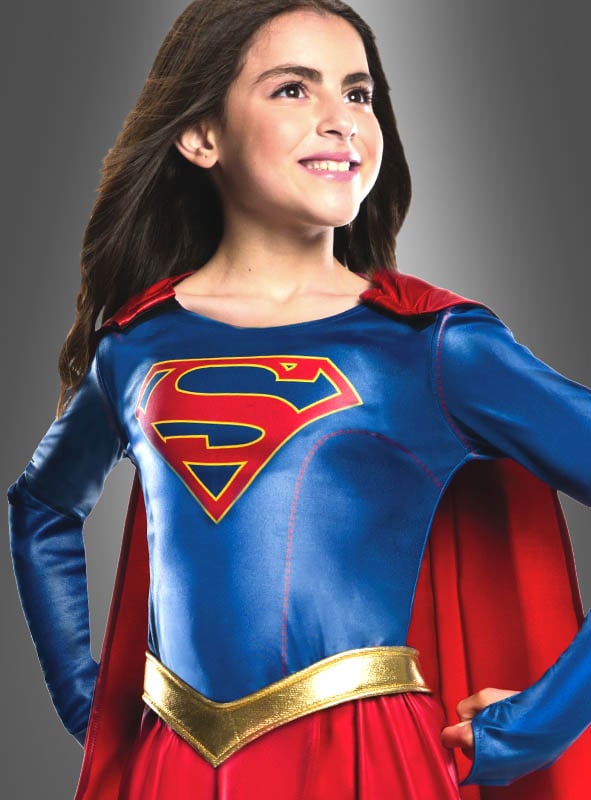 Ein lächelndes Kind steht mit den Händen in den Hüften da und trägt das Supergirl-Kostüm für Kinder – ein offiziell lizenziertes DC-Comics-Kostüm mit einem blau-roten Design, einem S-Emblem auf der Brust und einem roten Umhang –, das perfekt für jeden jungen Superhelden-Fan ist.