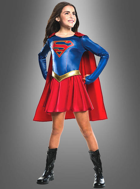 Ein junges Mädchen posiert selbstbewusst in einem Supergirl-Kostüm für Kinder, bestehend aus einem blauen Oberteil, einem roten Rock und einem Umhang, schwarzen Stiefeln und einem goldenen Gürtel – einem originalen Lizenzkostüm von DC Comics – vor einem grauen Hintergrund.
