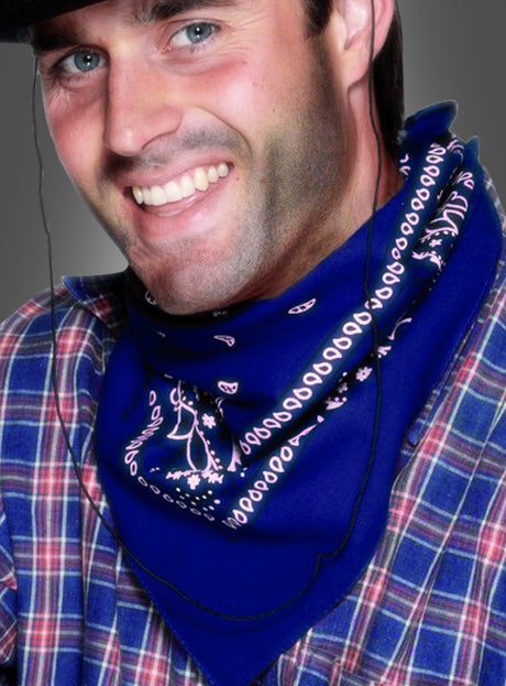 Eine lächelnde Person, die einen dunklen Hut, ein blaues Cowboy-Bandana mit weißen Mustern und ein kariertes Hemd trägt, wird vor einem grauen Hintergrund fotografiert.