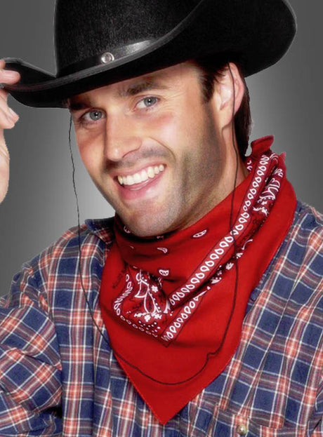 Ein lächelnder Mann in einem karierten Hemd, einem roten Cowboy-Bandana und einem schwarzen Cowboyhut zieht den Hut, während er vor einem schlichten grauen Hintergrund posiert.