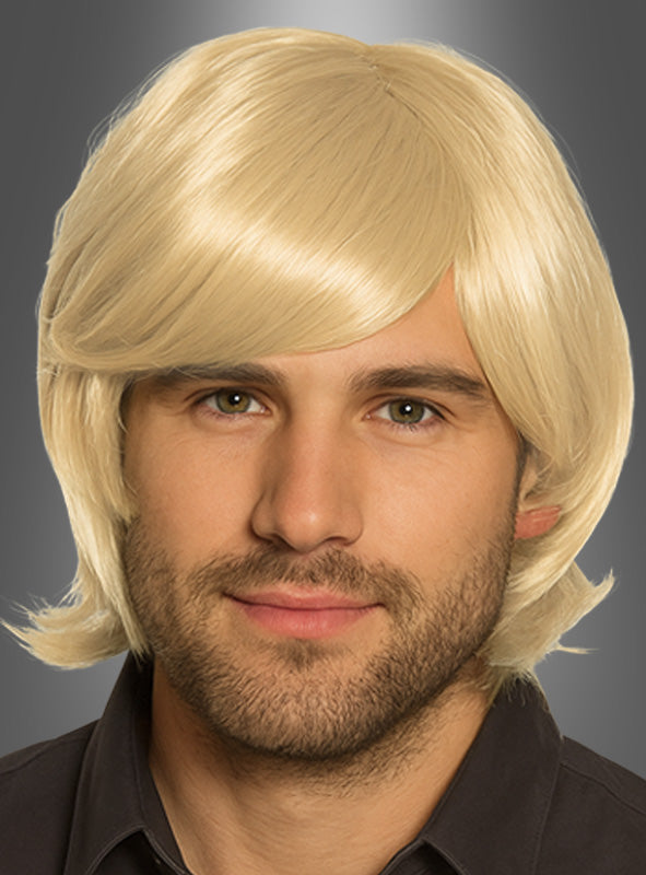 Ein Mann mit heller Haut und Bart, der eine kurze, blonde Howard-Perücke im Stil der 70er Jahre und ein dunkles Hemd mit Kragen trägt und vor einem grauen Hintergrund mit Farbverlauf nach vorne blickt.