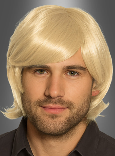 Ein Mann mit heller Haut und Bart, der eine kurze, blonde Howard-Perücke im Stil der 70er Jahre und ein dunkles Hemd mit Kragen trägt und vor einem grauen Hintergrund mit Farbverlauf nach vorne blickt.