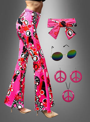 Eine Frau präsentiert die „Hippie-Schlaghose“ mit Accessoires: dazu gehören trendige rosa und mehrfarbige Schlaghosen, kombiniert mit einem passenden Stirnband, einer runden Sonnenbrille und rosa Peace-Zeichen-Ohrringen – die ideale Wahl für jede Hippie-Mottoparty –, während sie vor einem grauen Hintergrund posiert.