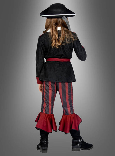 Ein Mädchen, das das „Cooles Piraten-Outfit für Mädchen“ trägt – bestehend aus einem schwarzen Hut, einer schwarzen Jacke, einer roten Schärpe, einer gestreiften Hose mit roten Rüschen und schwarzen Stiefeln –, steht mit dem Rücken zur Kamera vor einem grauen Hintergrund mit Farbverlauf.