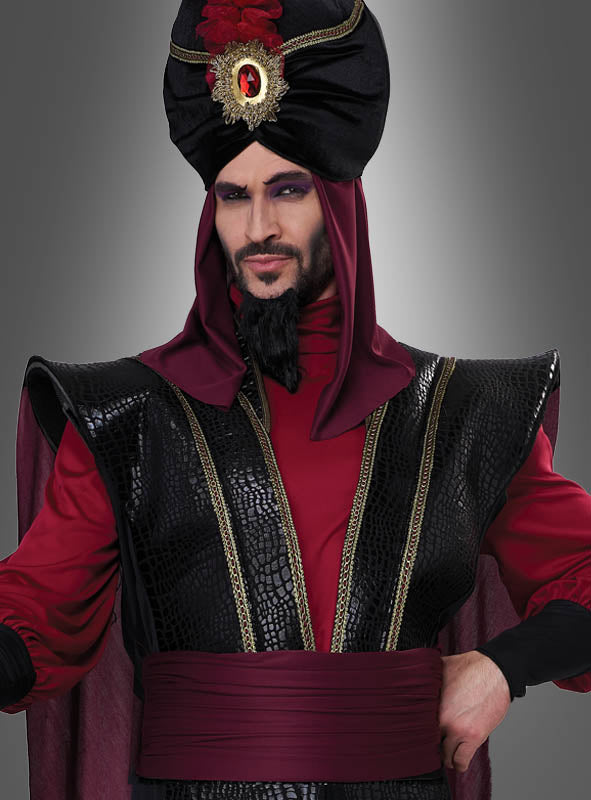 Ein Mann im „Düsterer Sultan“-Kostüm für Herren – eine auffällige rot-schwarze Robe mit kunstvoll verzierter Schulterpanzerung, einem hohen, mit Edelsteinen besetzten Turban und einem stilisierten Bart – posiert vor einem grauen Hintergrund. Damit ist dieses Kostüm die ideale Wahl für alle, die den geheimnisvollen Charme der Traditionen aus „1001 Nacht“ verkörpern möchten.